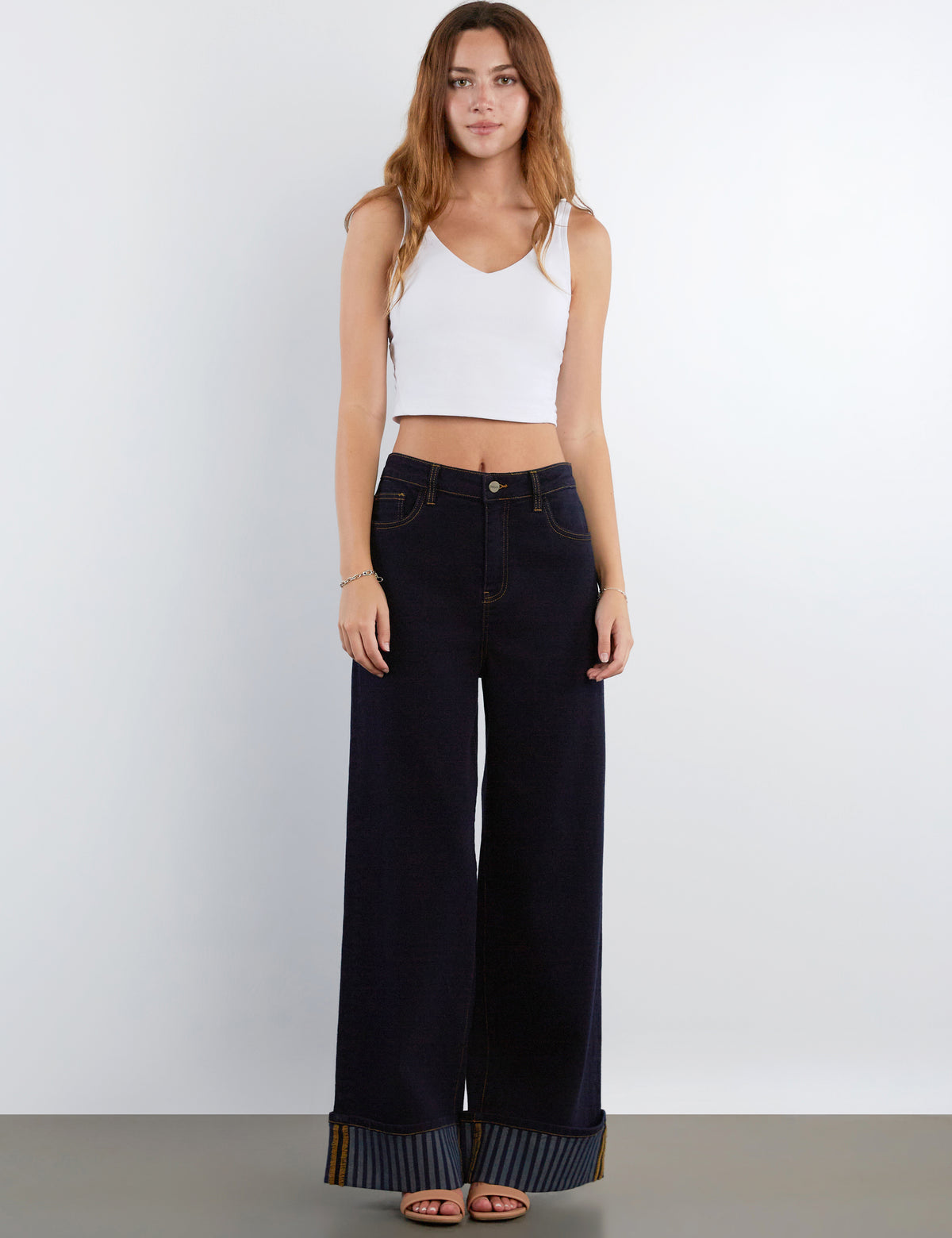 A woman modeling a pair of dark wide-leg jeans with striped turned-up hems, paired with a white crop top, standing against a light gray studio background.