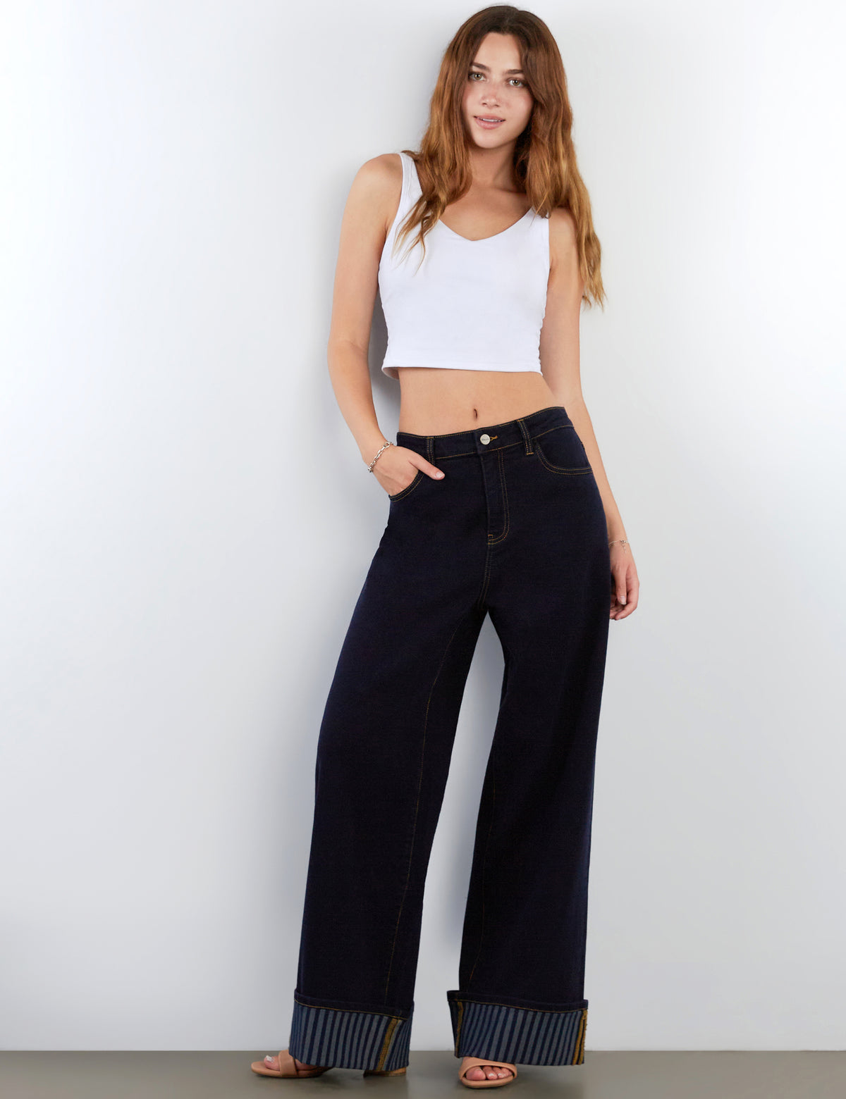 A woman modeling dark wide-leg jeans with a cuffed hem, paired with a white crop top, standing against a plain light gray wall.
