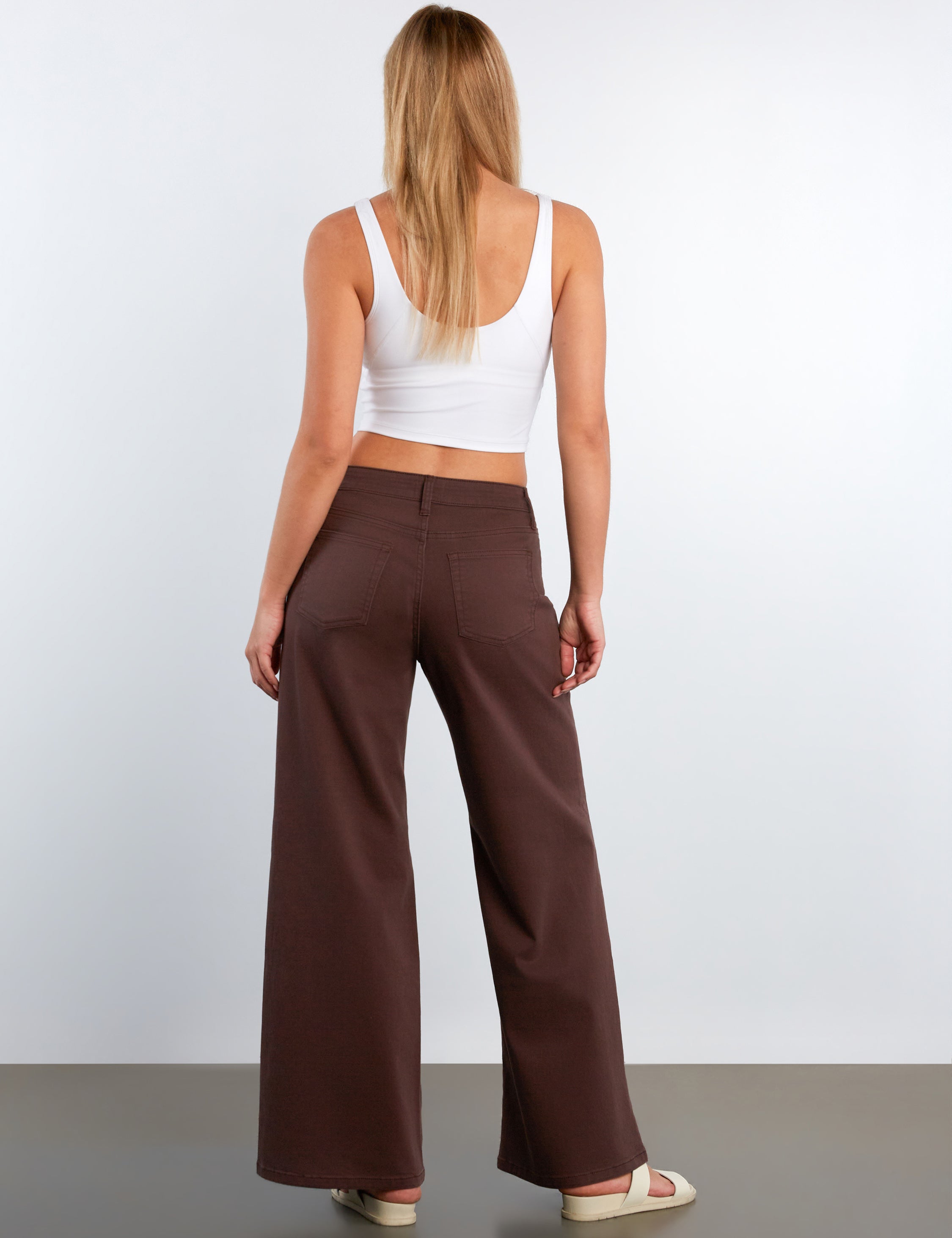 Woman wearing a white tank top and brown wide-leg pants against a plain background