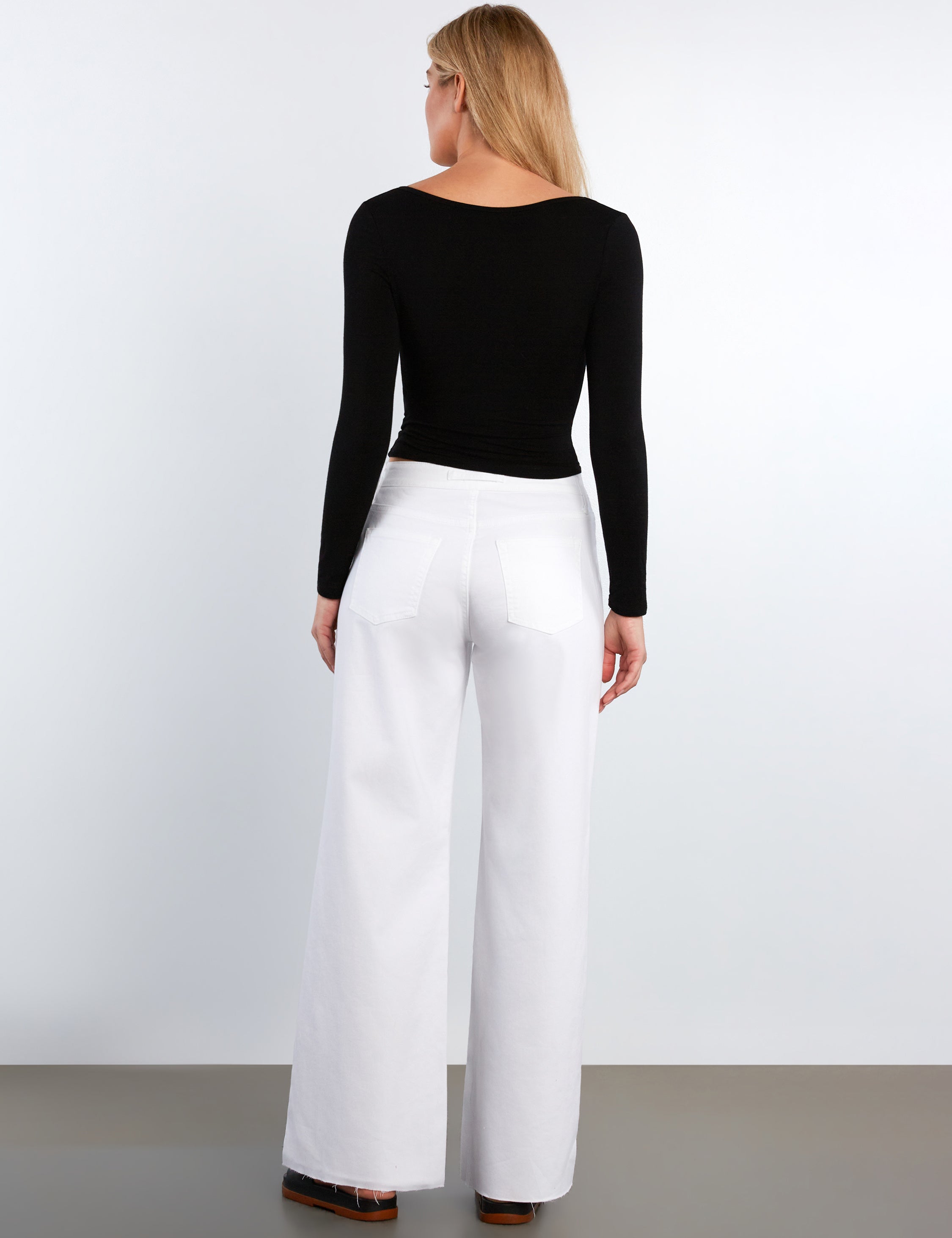 Woman wearing a black long-sleeve top and white pants against a white background