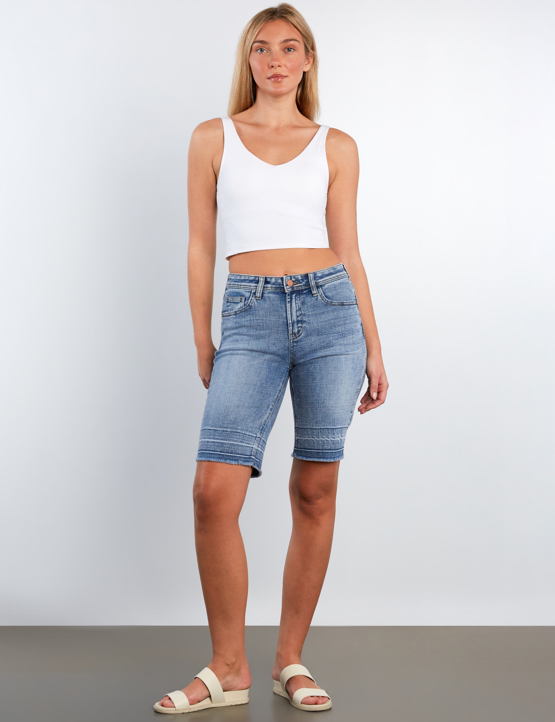 Woman wearing a white tank top and blue denim shorts against a plain background