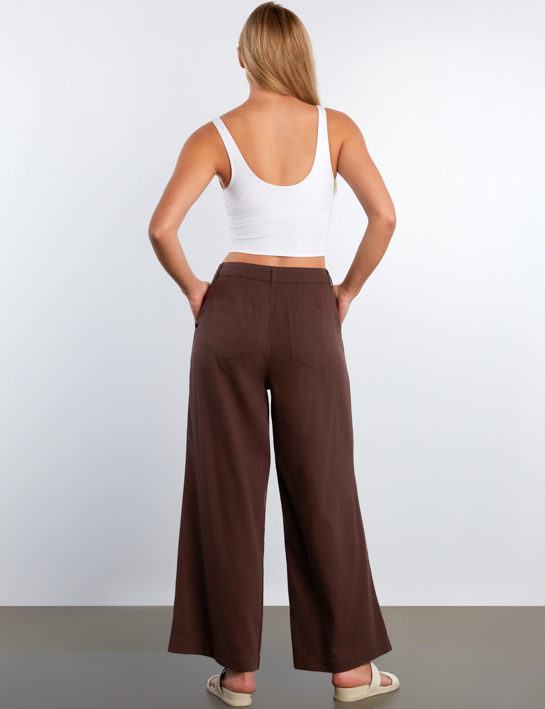 Woman wearing a white tank top and brown wide-leg pants against a plain background