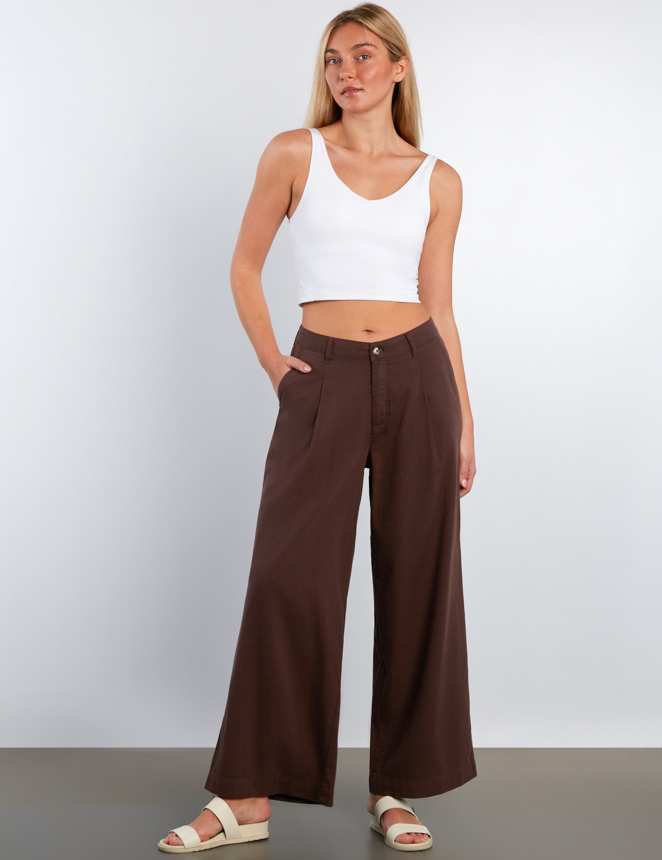 Woman wearing a white tank top and brown wide-leg pants against a plain background