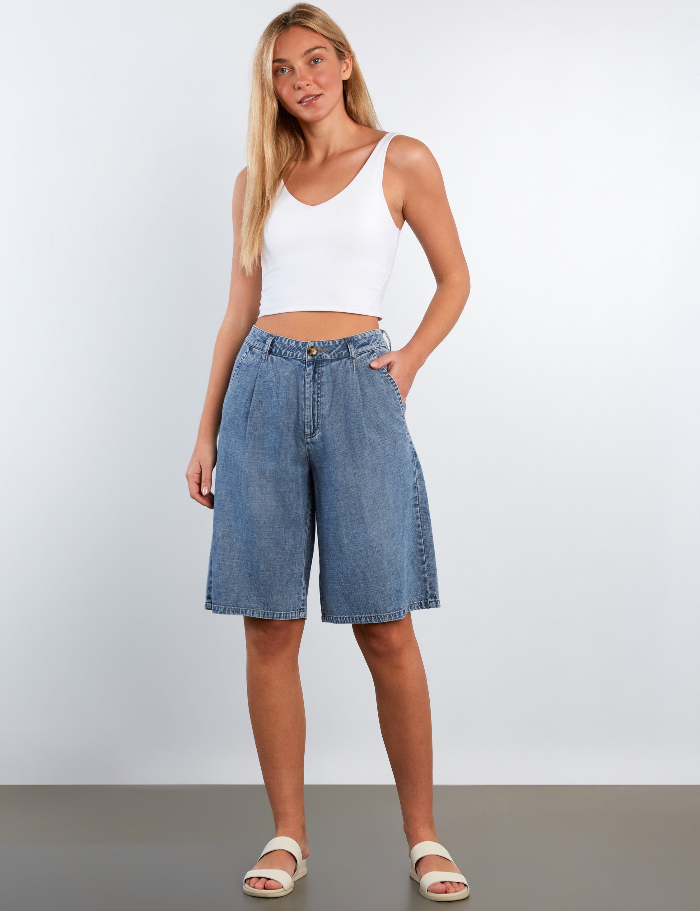 Woman wearing a white tank top and blue denim shorts against a plain background
