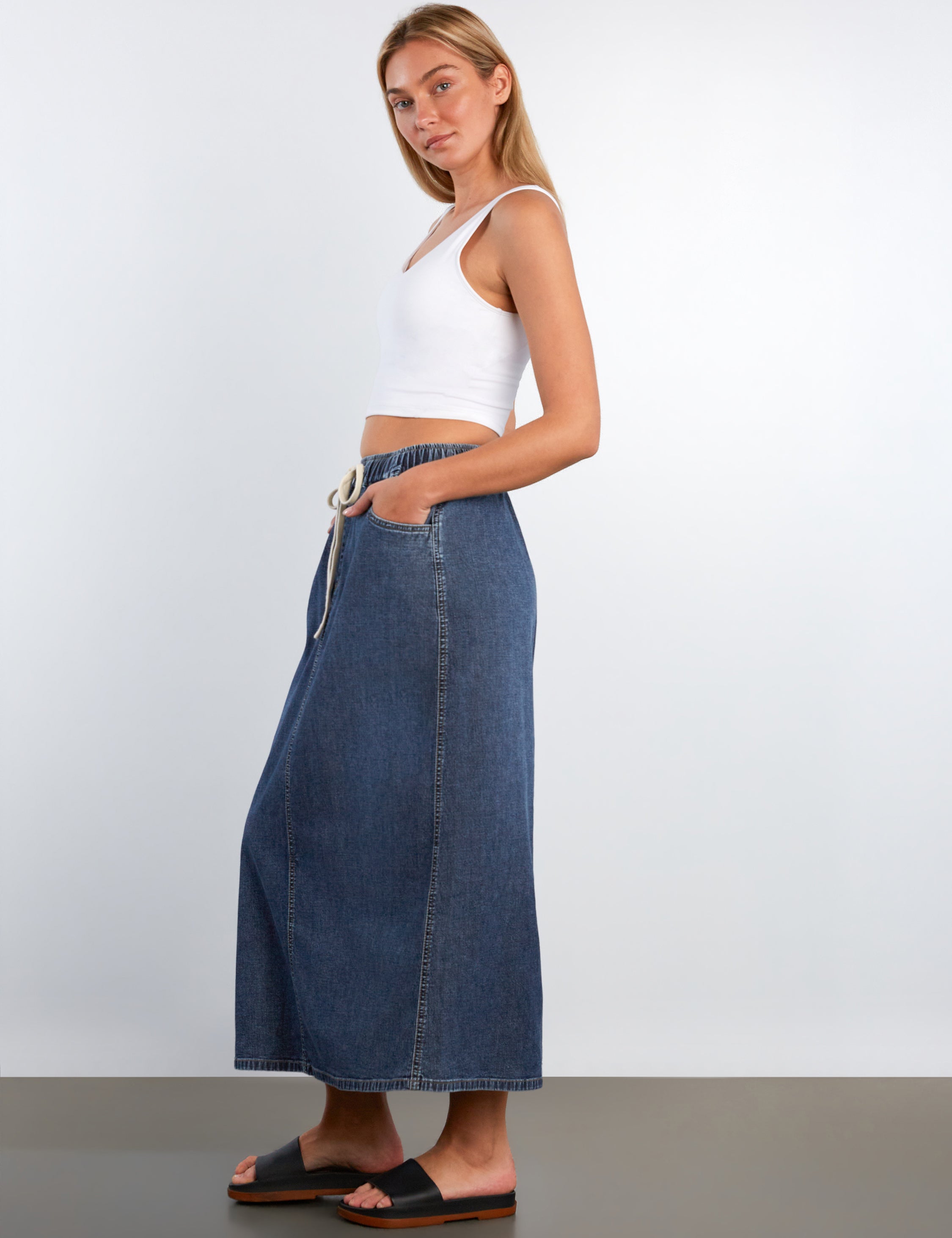 Woman wearing a white crop top and long denim skirt against a plain background
