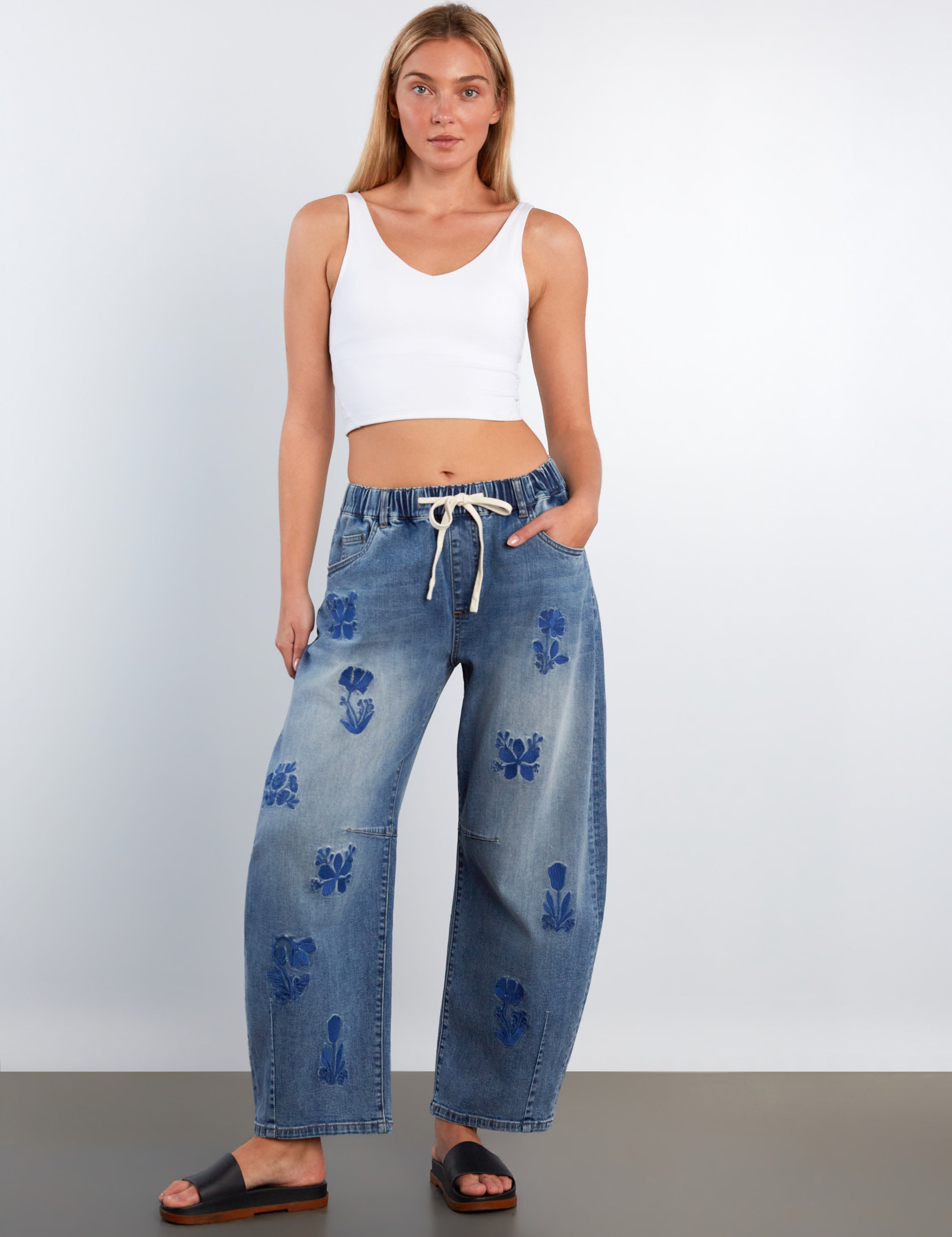 Woman wearing blue jeans with floral patterns and a white top on a plain background