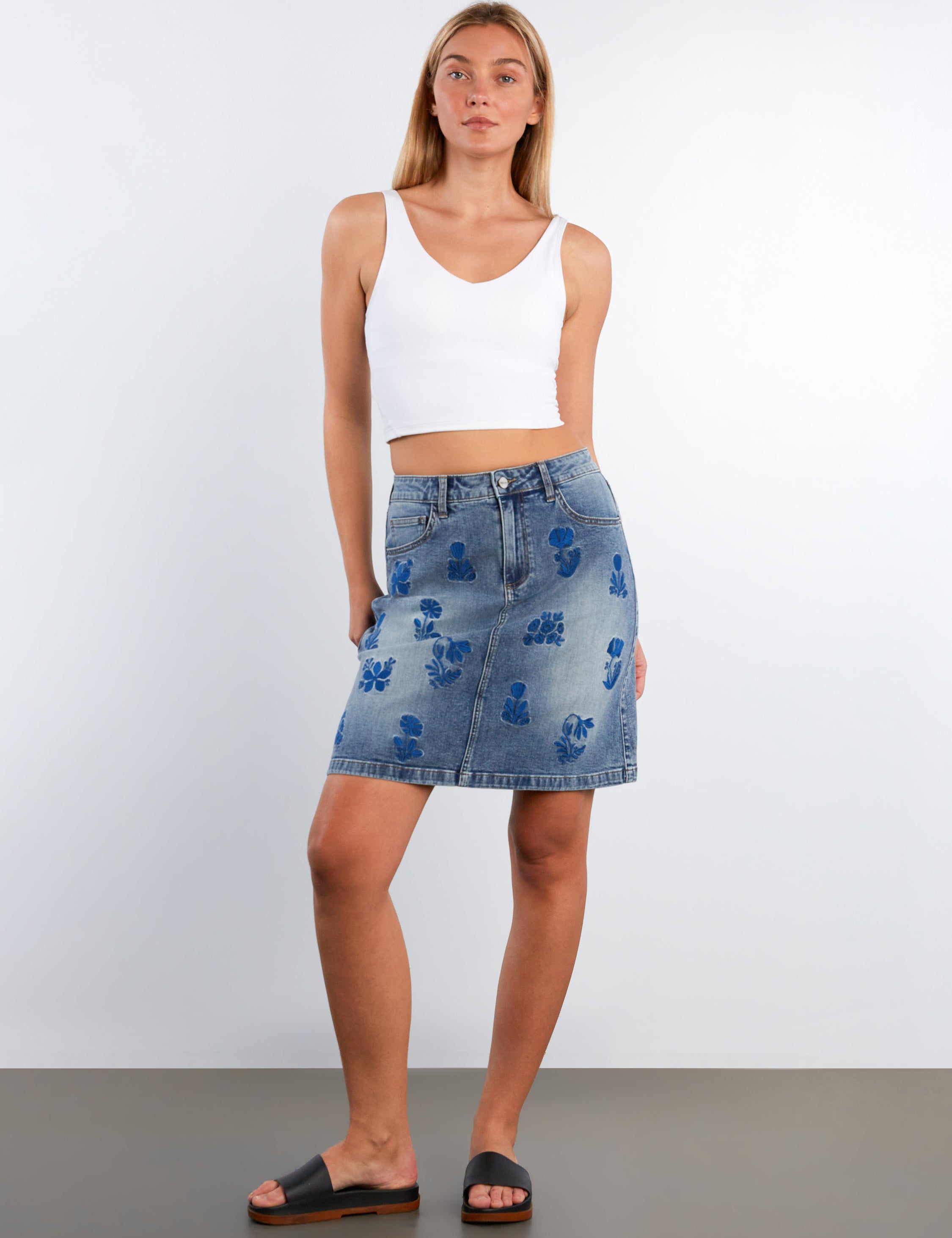 Woman wearing a white tank top and blue floral denim skirt on a plain background
