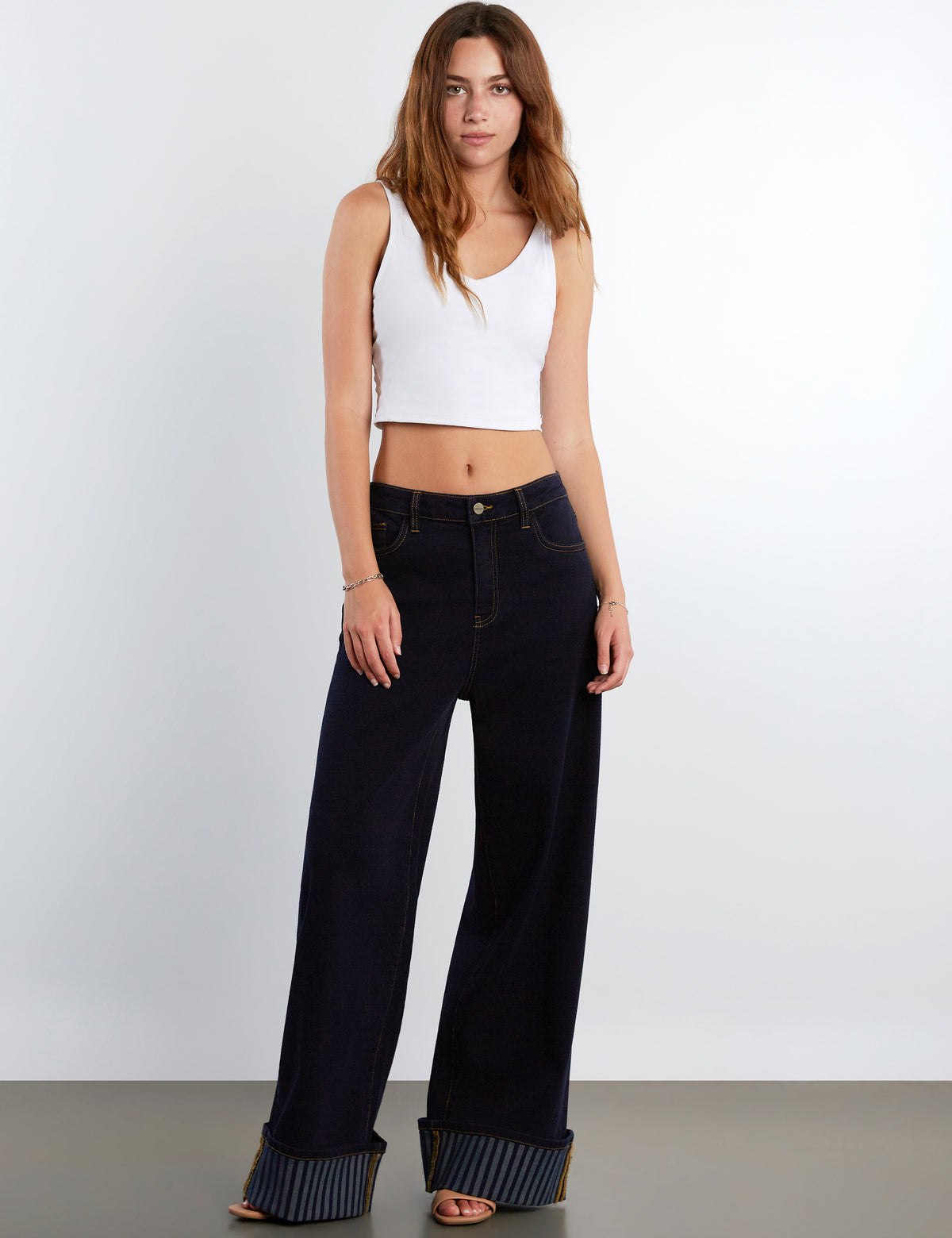 A woman modeling a pair of dark wide-leg jeans with striped turned-up hems, paired with a white ribbed crop top, against a light gray studio background.