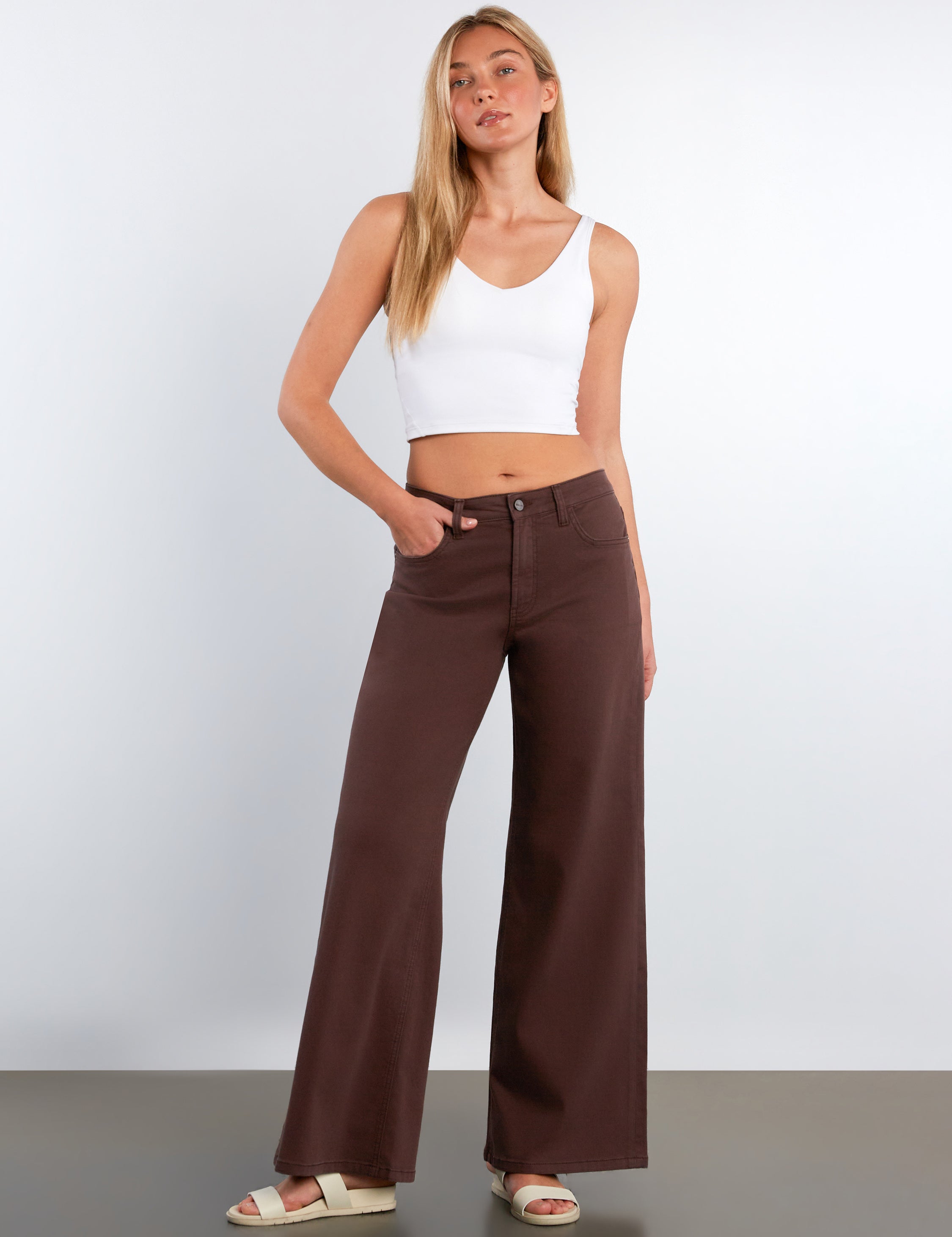 Woman wearing a white tank top and brown wide-leg pants against a plain background