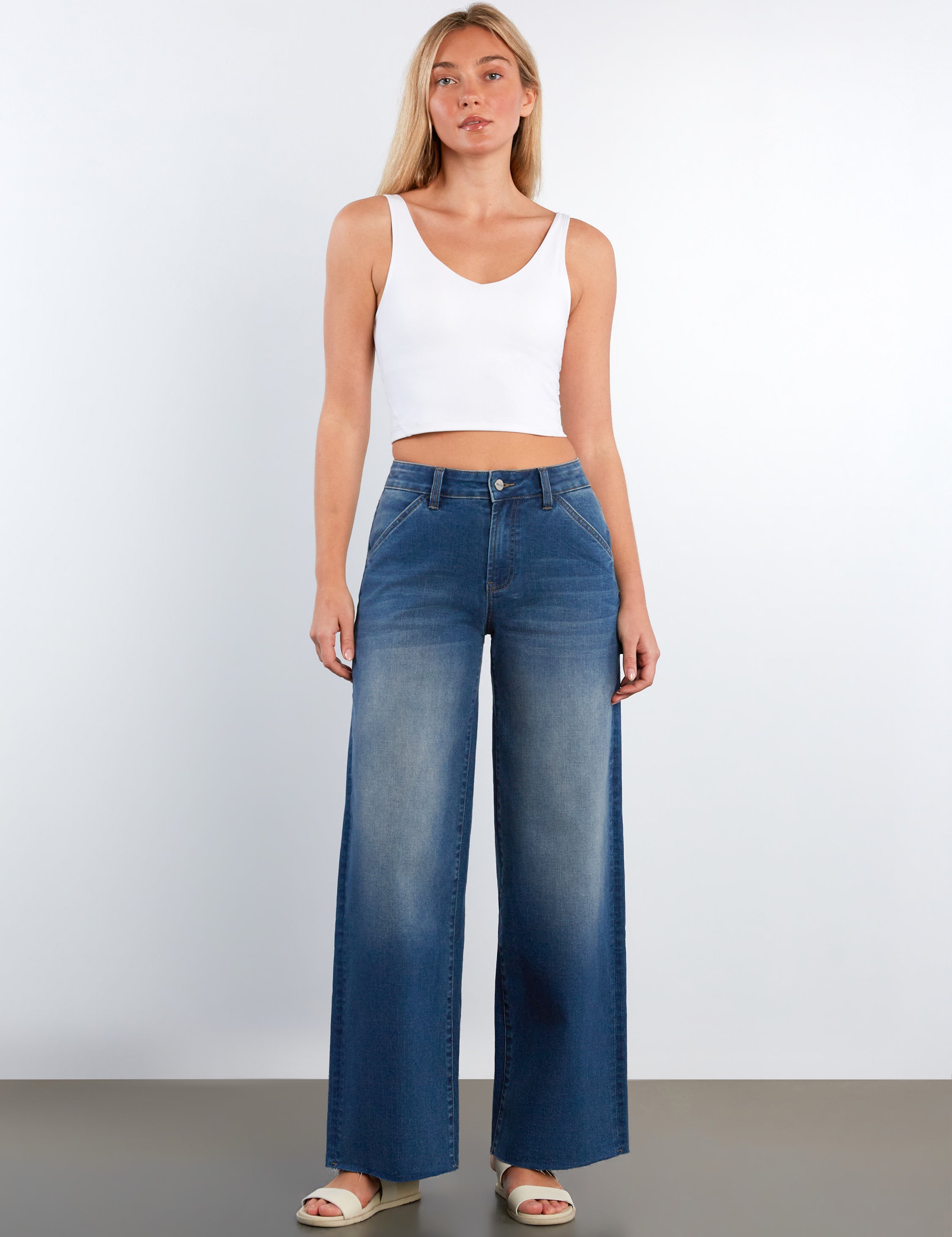 Woman wearing blue wide-leg jeans and a white tank top against a plain background
