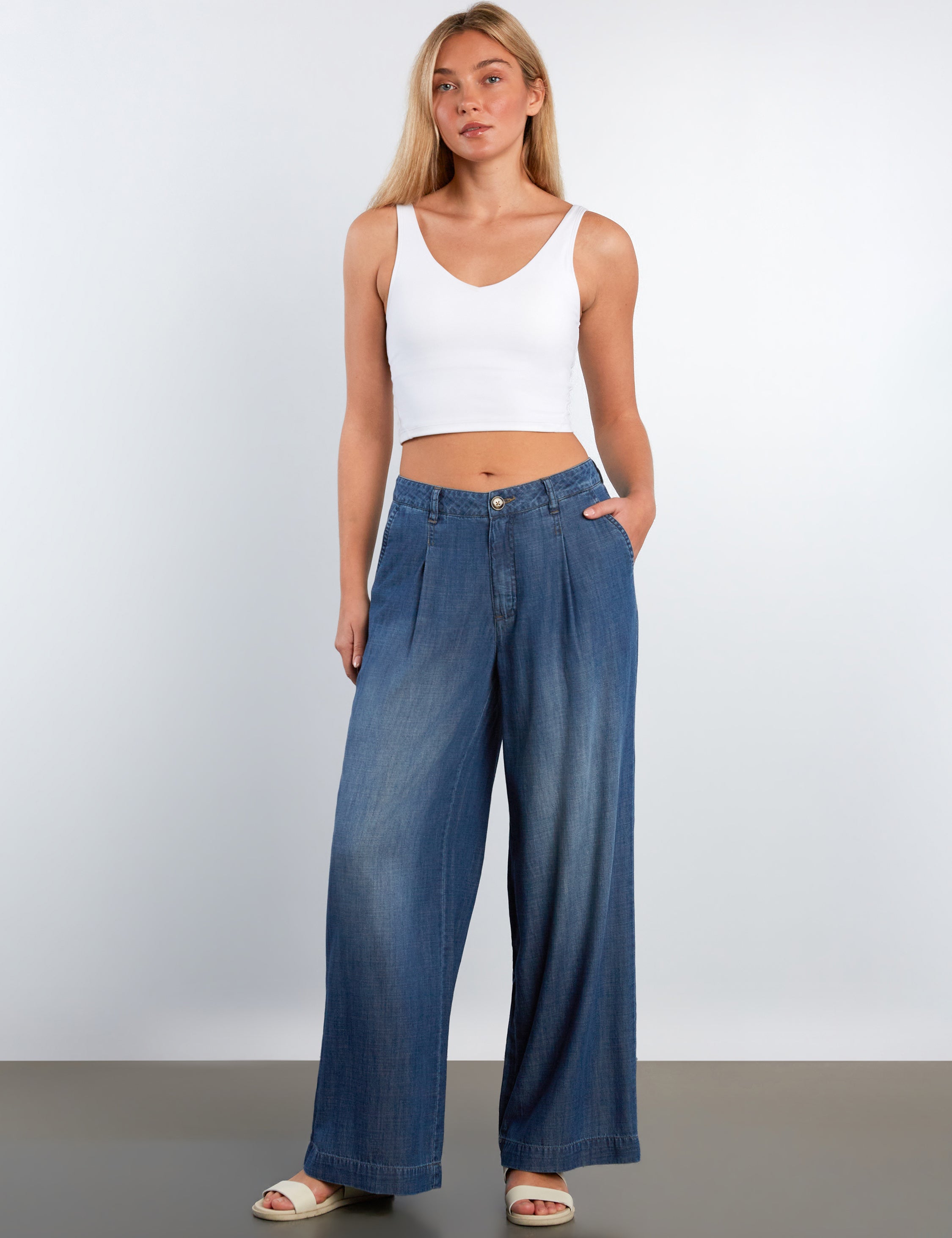 Woman wearing a white tank top and blue wide-leg jeans against a plain background
