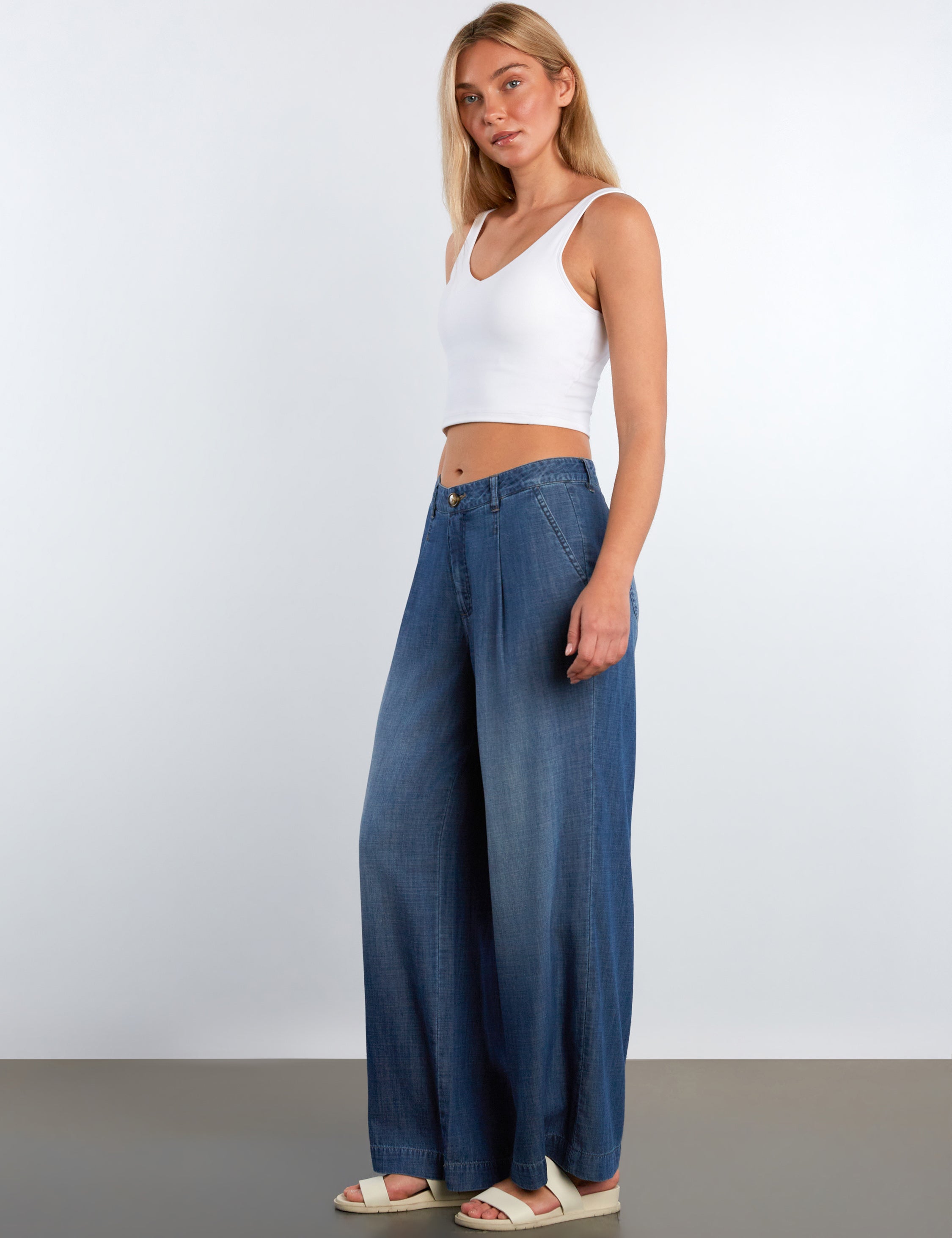 Woman wearing a white tank top and blue wide-leg jeans against a plain background