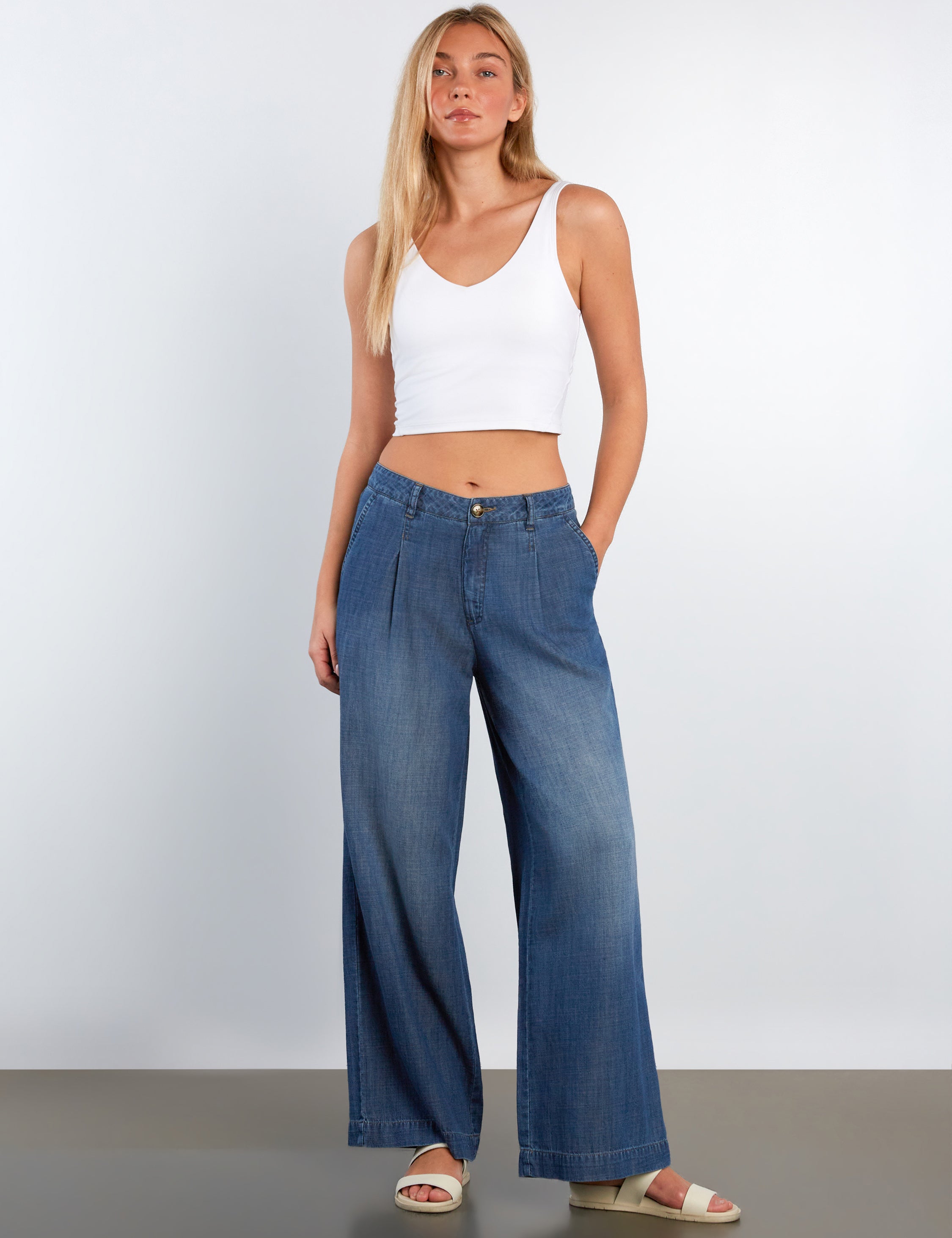 Woman wearing a white tank top and blue wide-leg jeans on a plain background