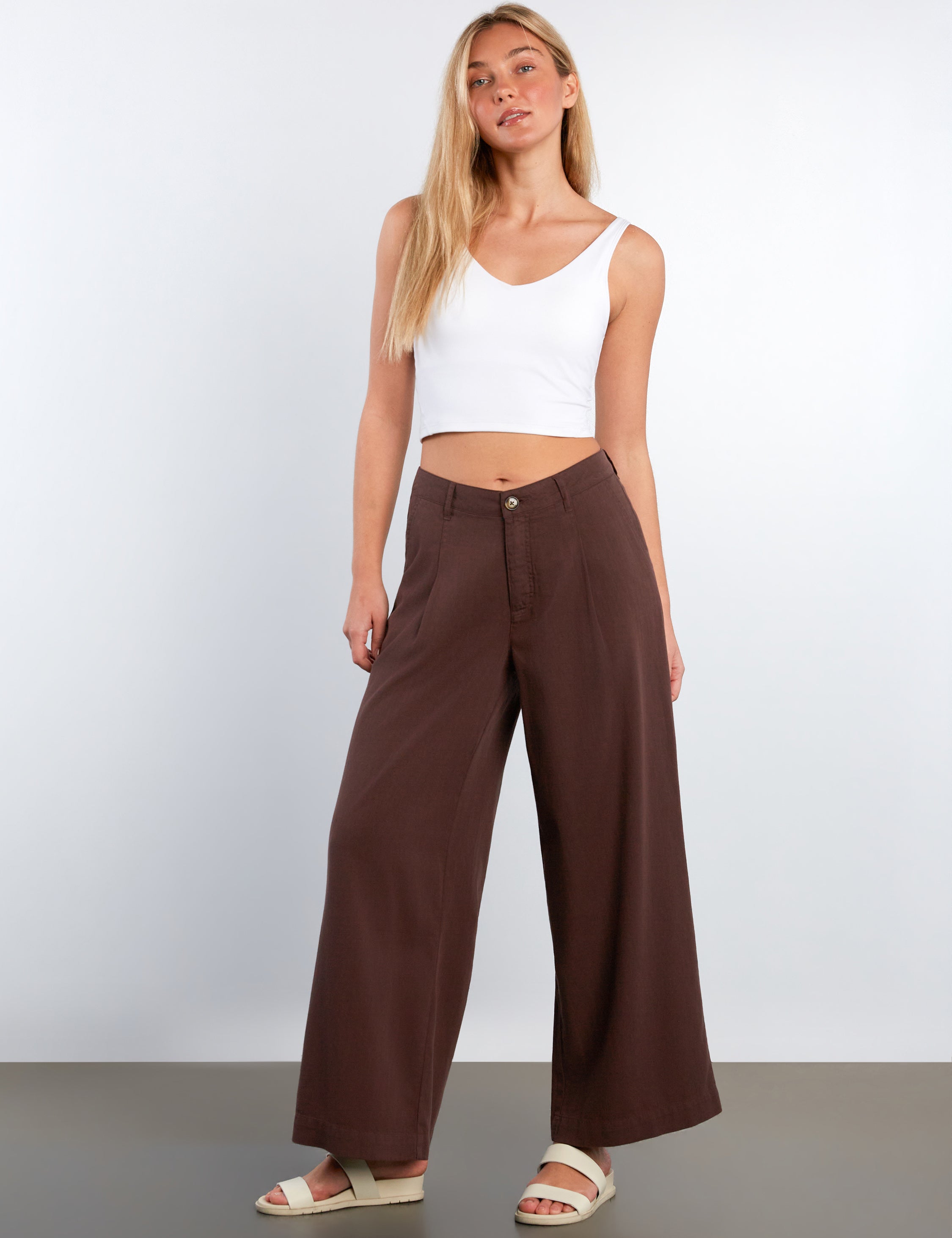 Woman wearing a white tank top and brown wide-leg pants against a plain background