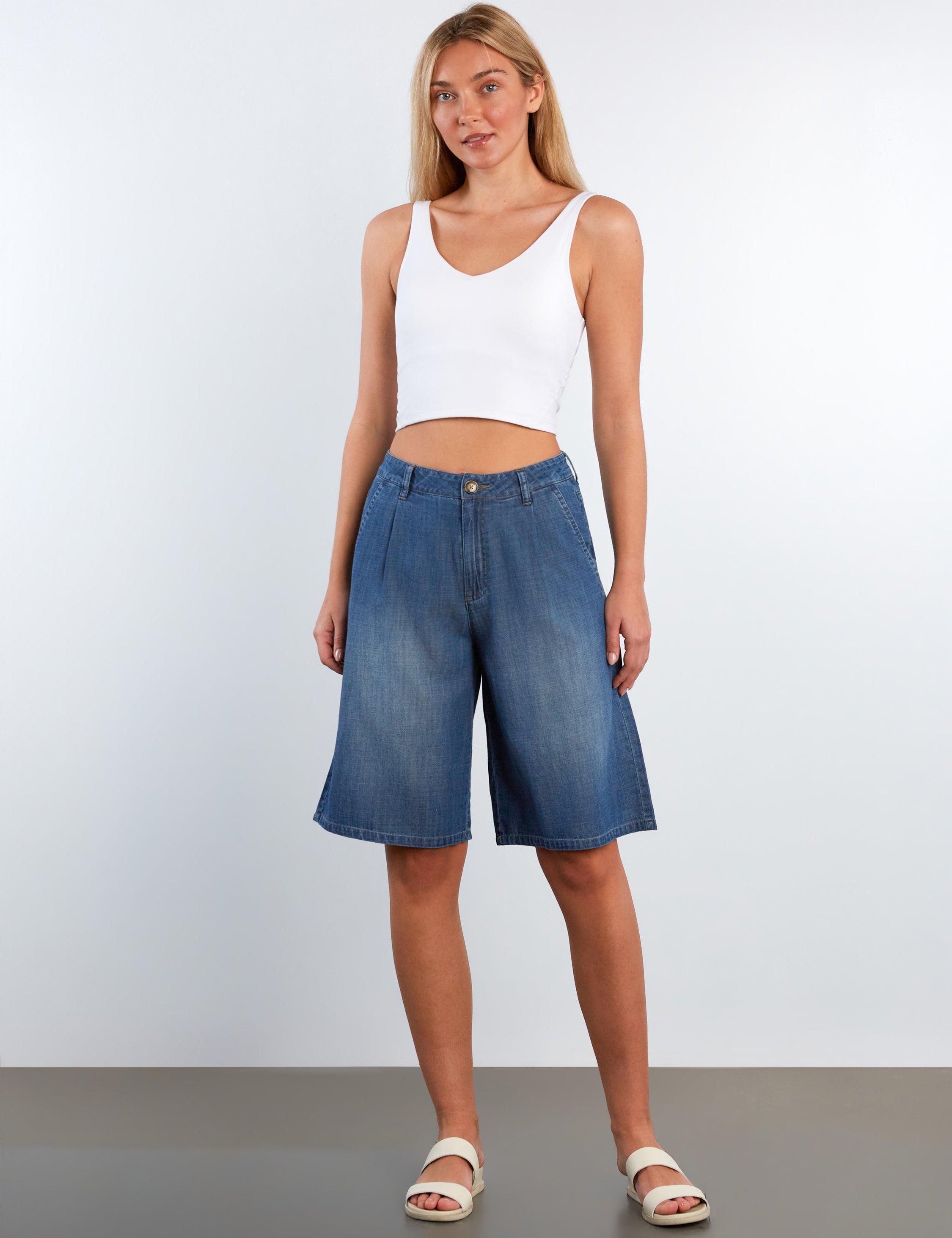 Woman wearing a white tank top and blue denim shorts against a plain background