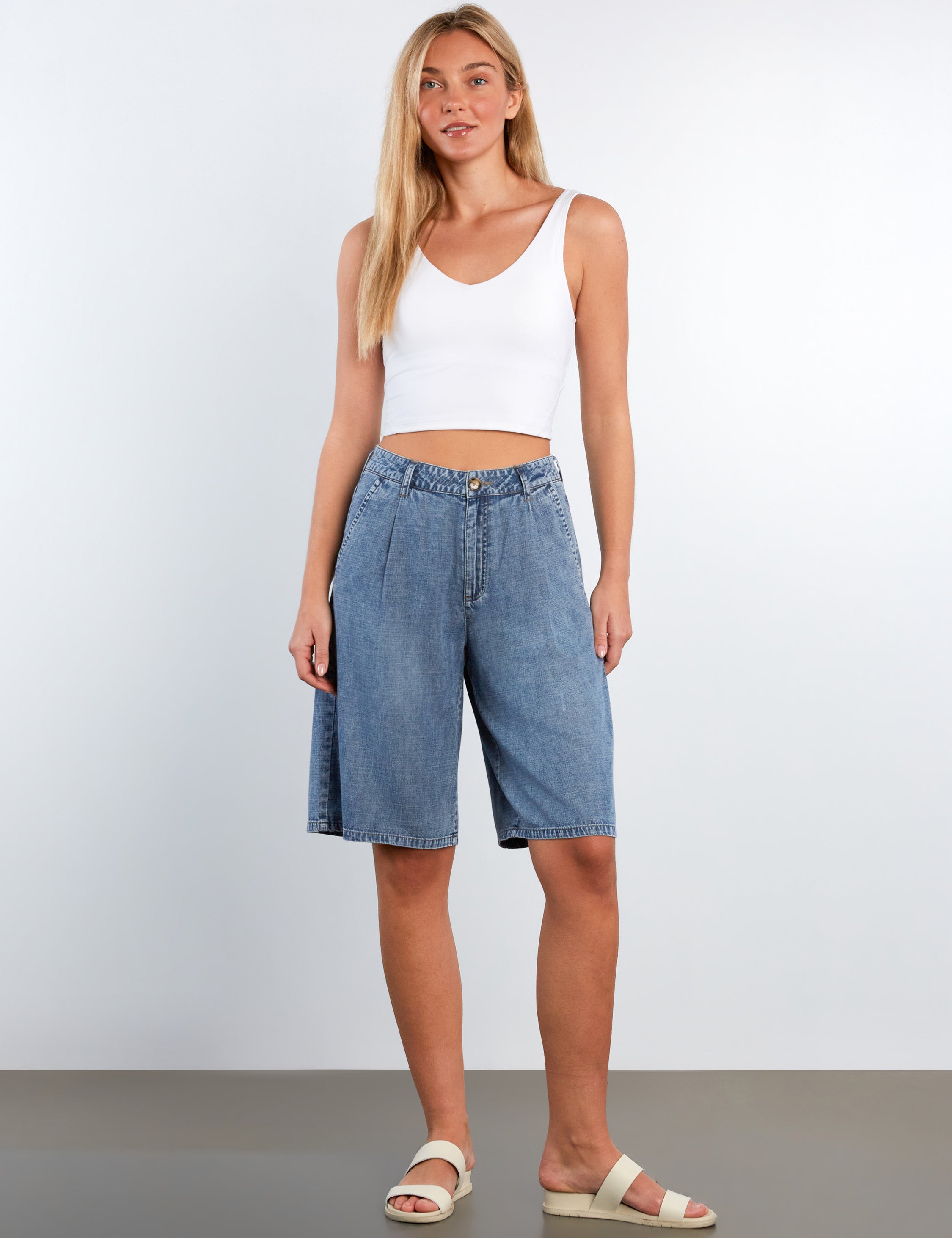 Woman wearing a white tank top and blue denim shorts on a plain background