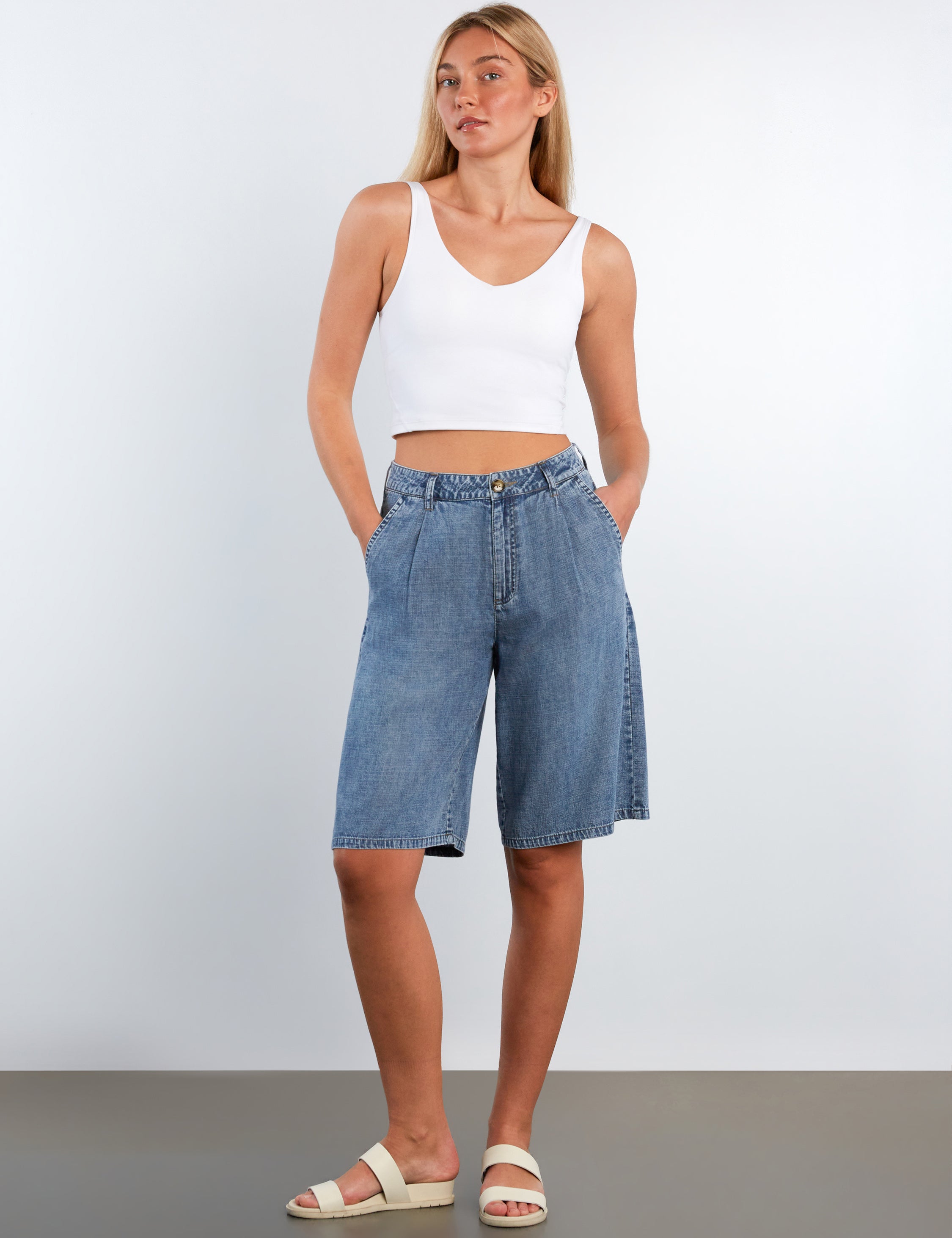 Woman wearing a white tank top and blue denim shorts against a plain background