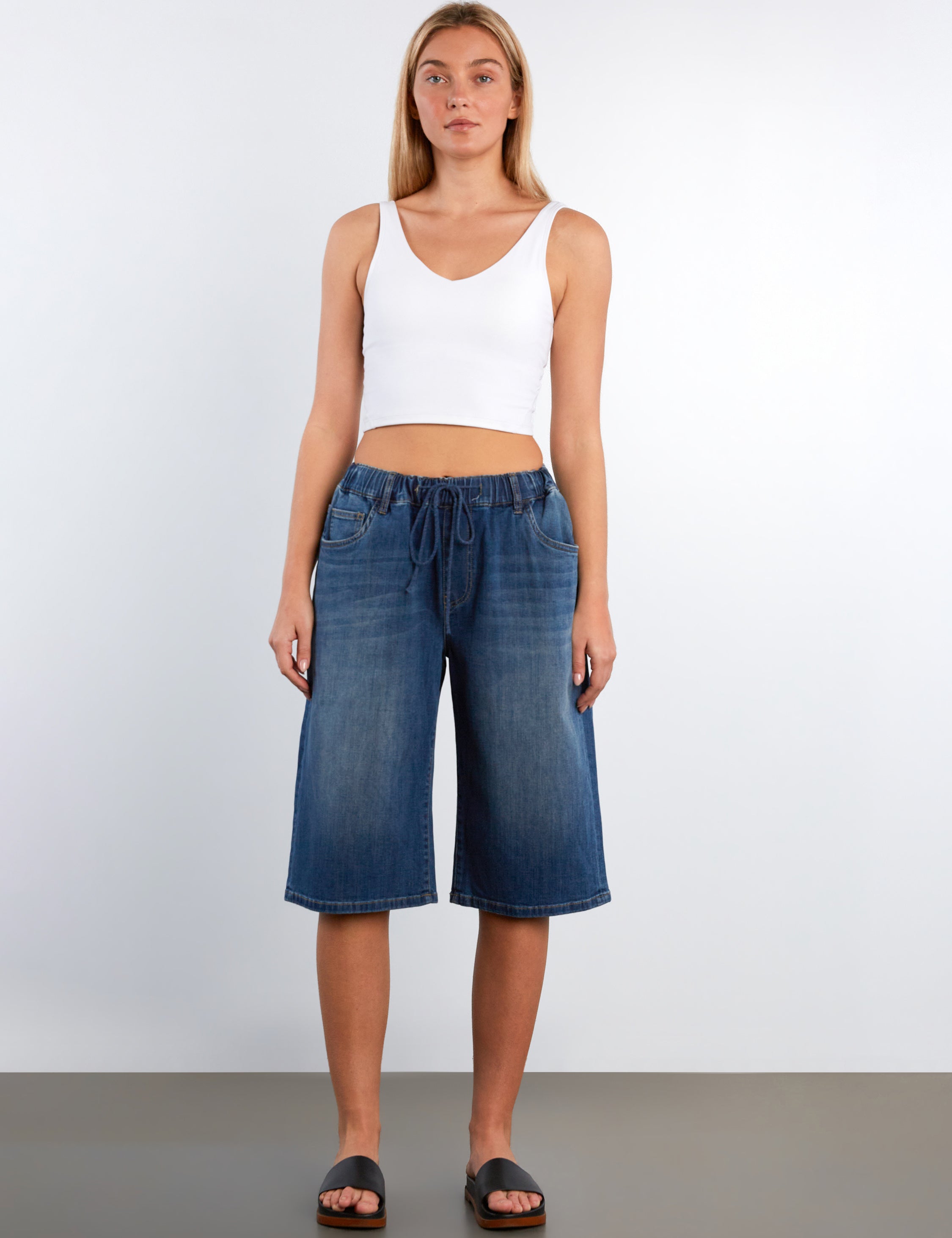 Woman wearing a white tank top and blue denim shorts against a plain background