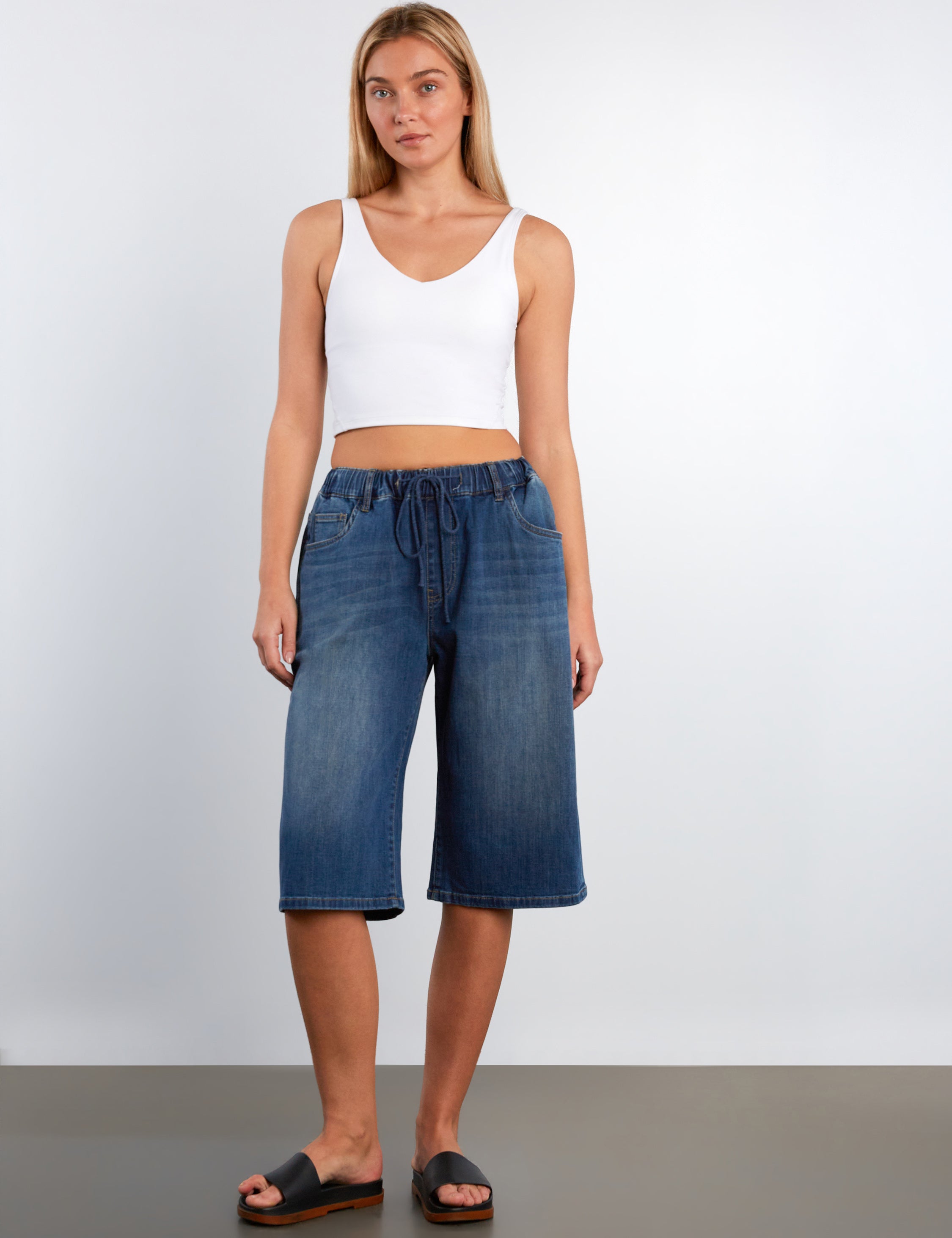Woman wearing a white tank top and blue denim shorts against a plain background
