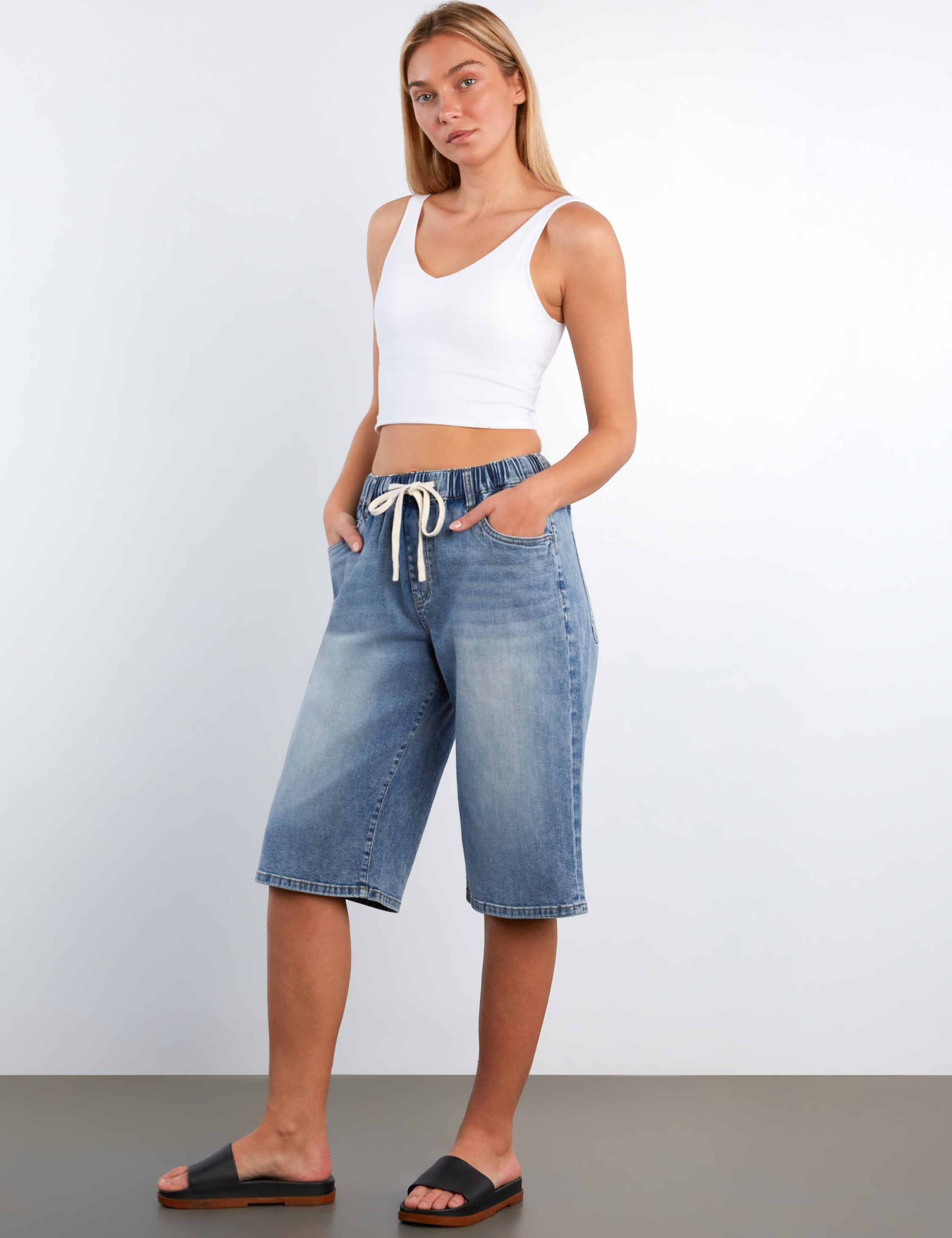 Woman wearing a white tank top and blue denim shorts against a plain background