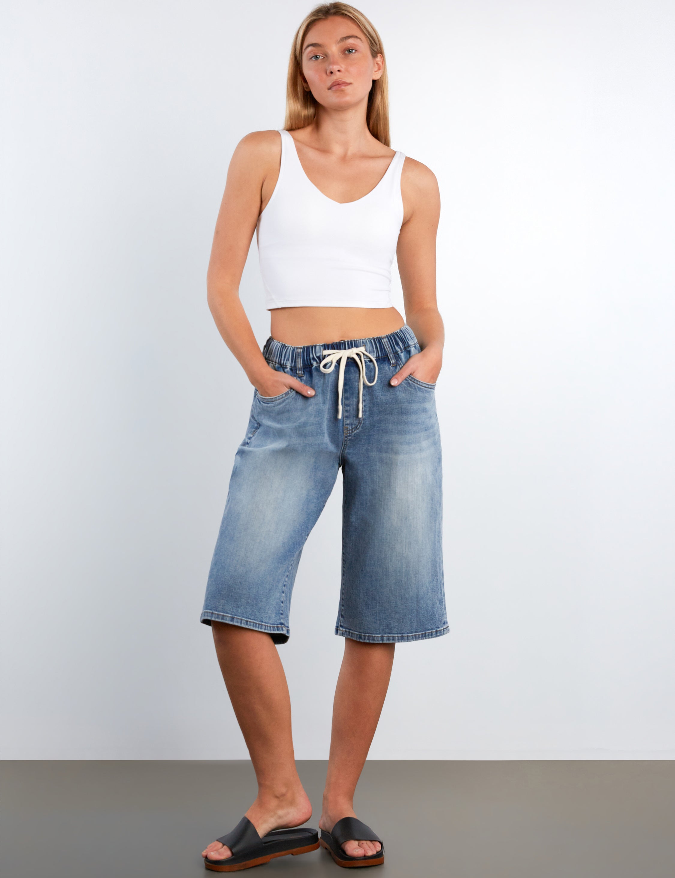 Woman wearing a white tank top and blue denim shorts against a plain background