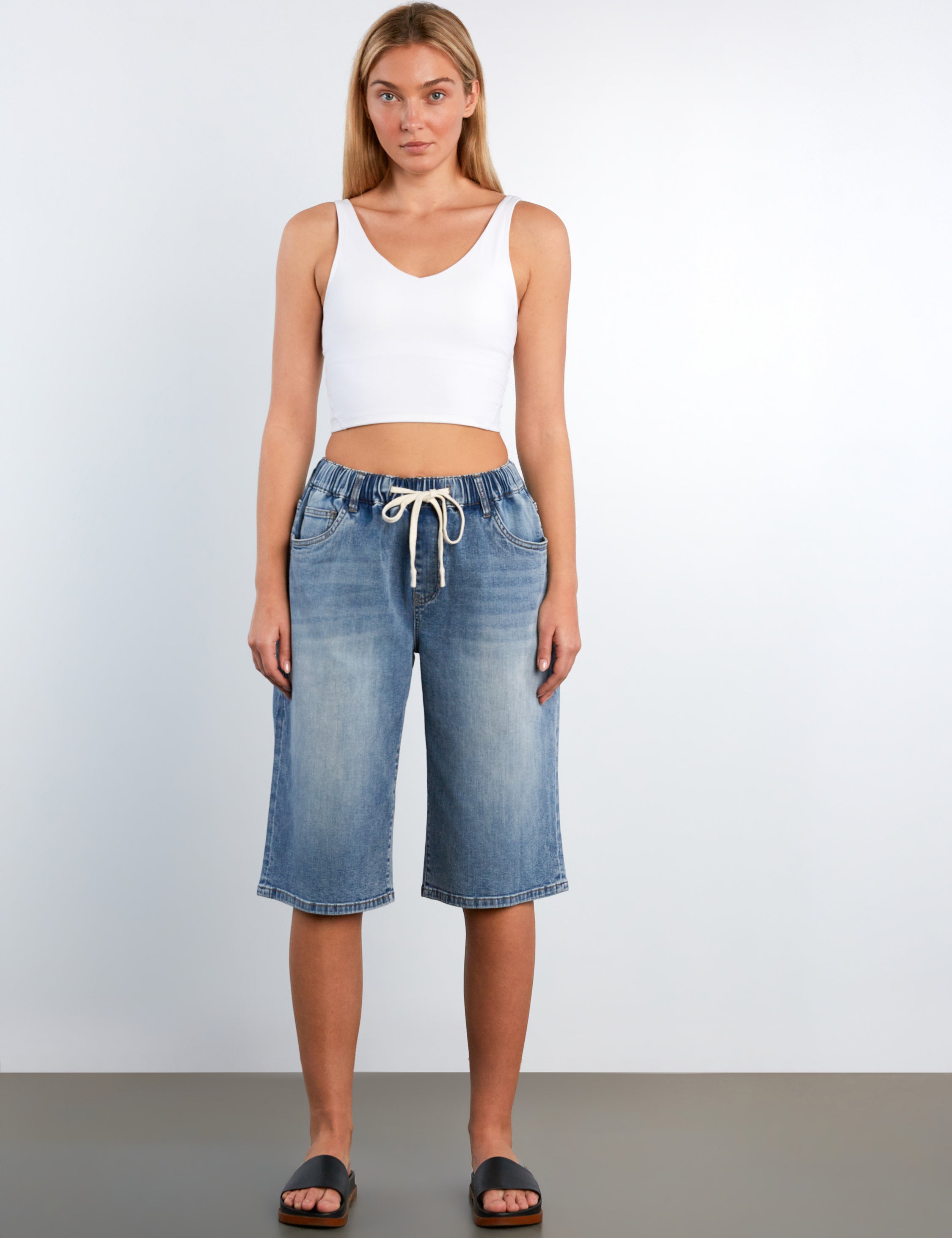 Woman wearing a white tank top and blue denim shorts on a plain background