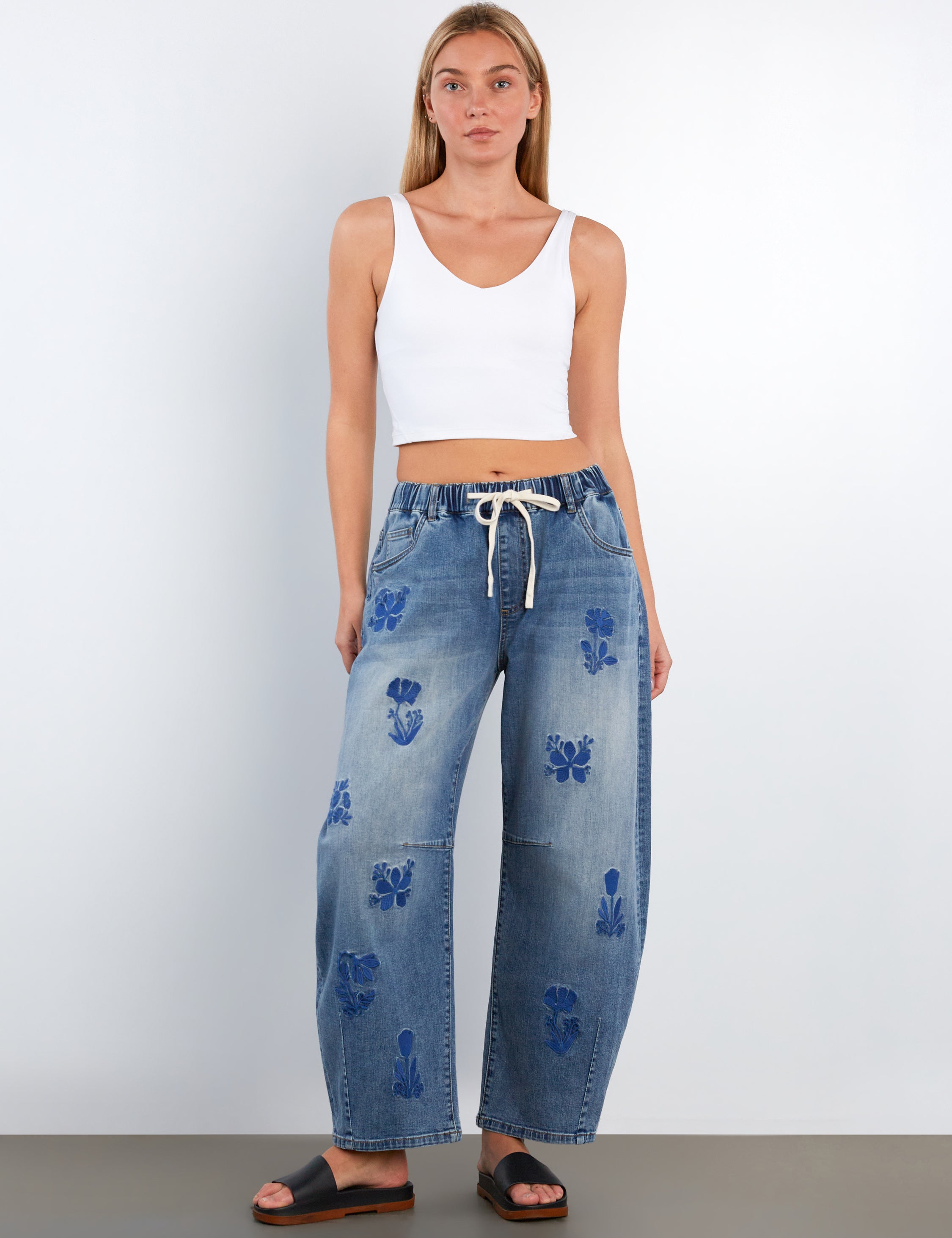 Woman wearing blue jeans with floral patterns and a white top on a plain background