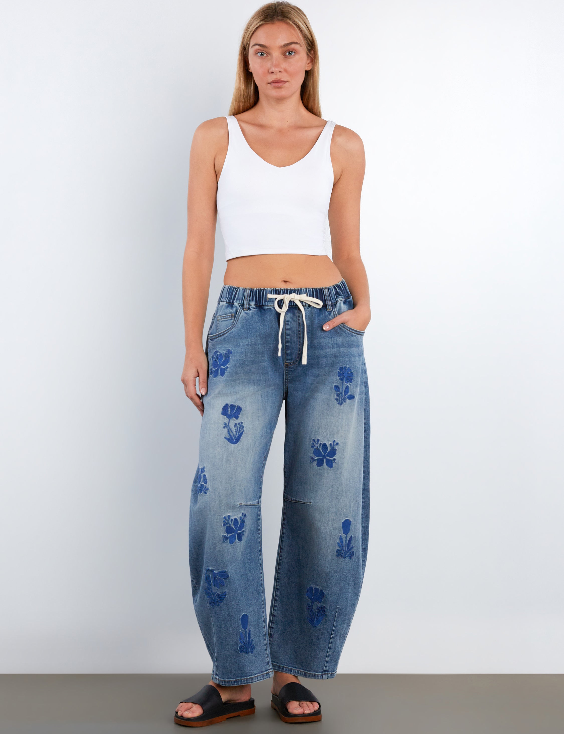 Woman wearing blue floral jeans and a white tank top on a plain background