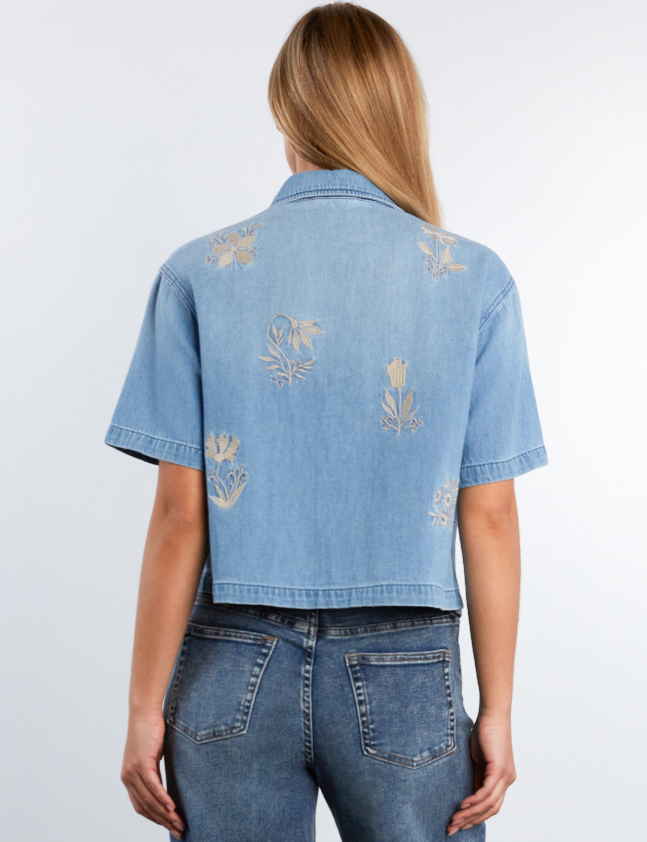 Person wearing a light blue denim shirt with floral embroidery on a white background