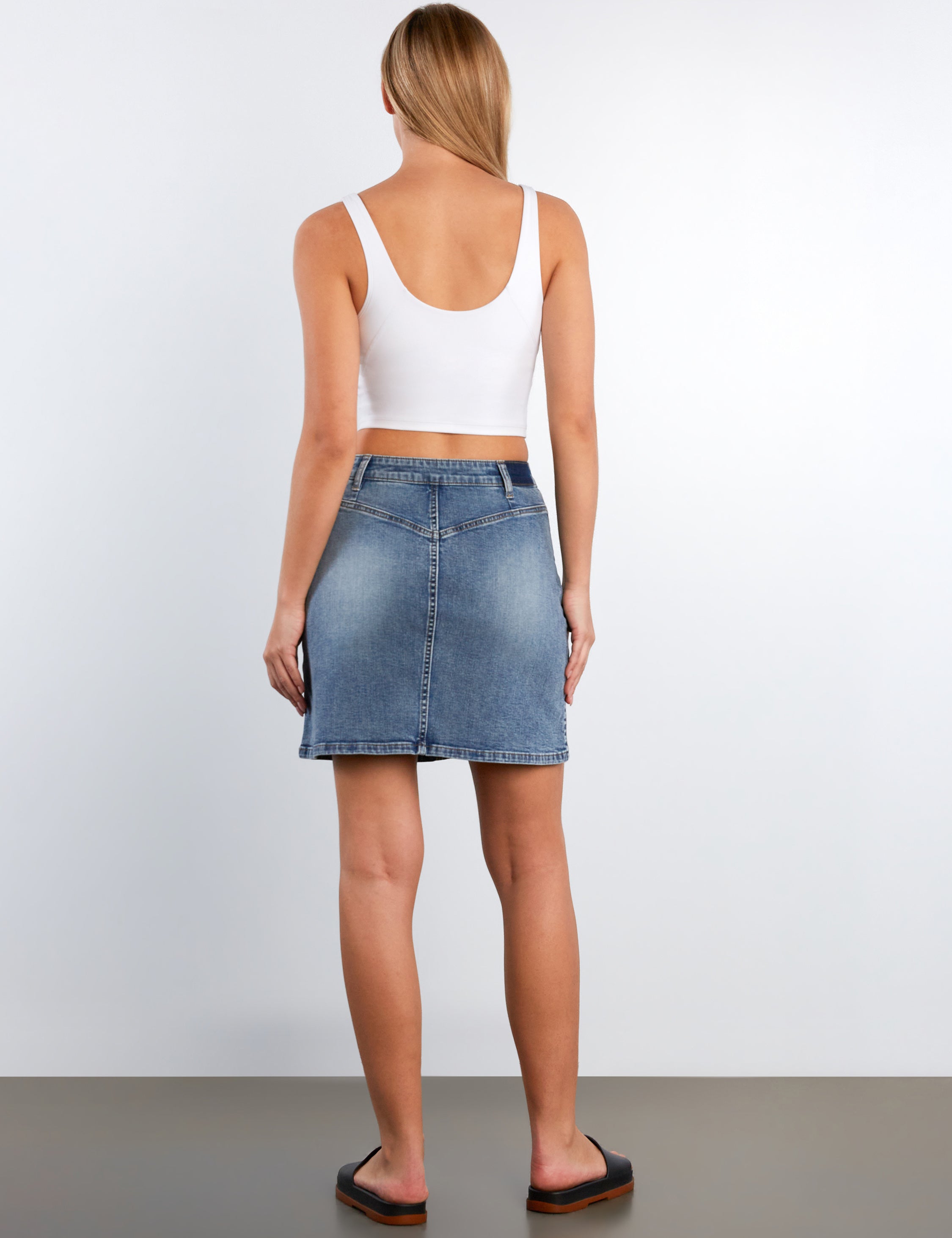 Person wearing a white tank top and blue denim skirt on a plain background
