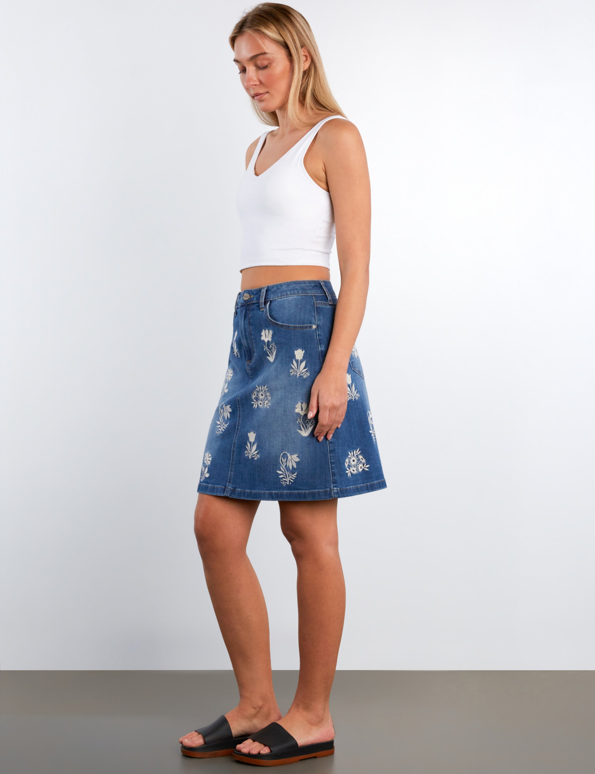 Woman wearing a white top and blue denim skirt with floral patterns on a plain background
